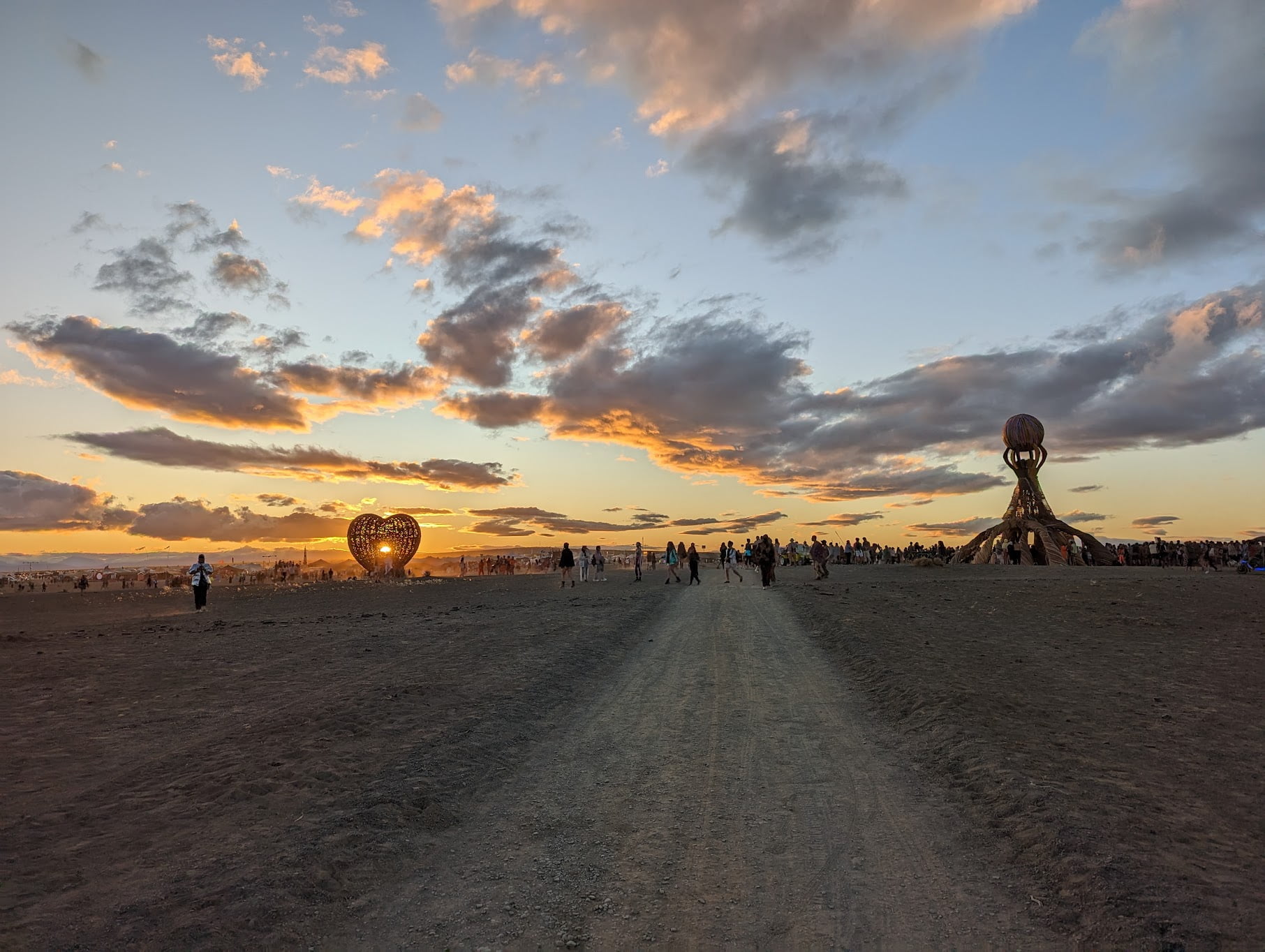 Geen kater meer na je festival? Ontdek de kracht van NMN Een breed onverhard pad leidt naar een druk festivalterrein in een woestijn, met veel mensen verspreid onder een lucht vol dramatische wolken bij zonsondergang. Sculpturen, waaronder een hart en een groot bouwwerk met een verhoogde bol, steken prominent af tegen de kleurrijke lucht.